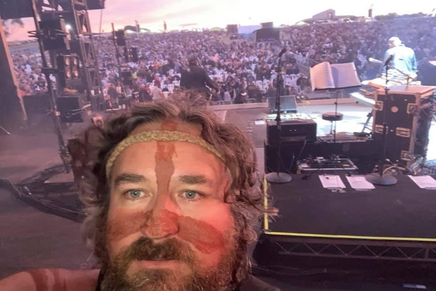 An Indigenous man with face paint takes a selfie overlooking the audience at a music festival