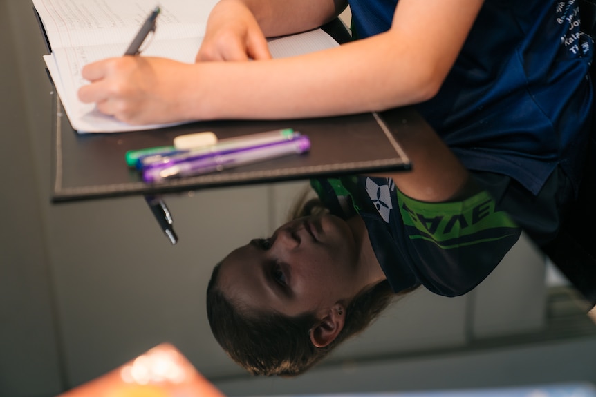 A reflection in a table of a young girl writing in a notepad, with pens, the notepad and her arm visible above her reflection.