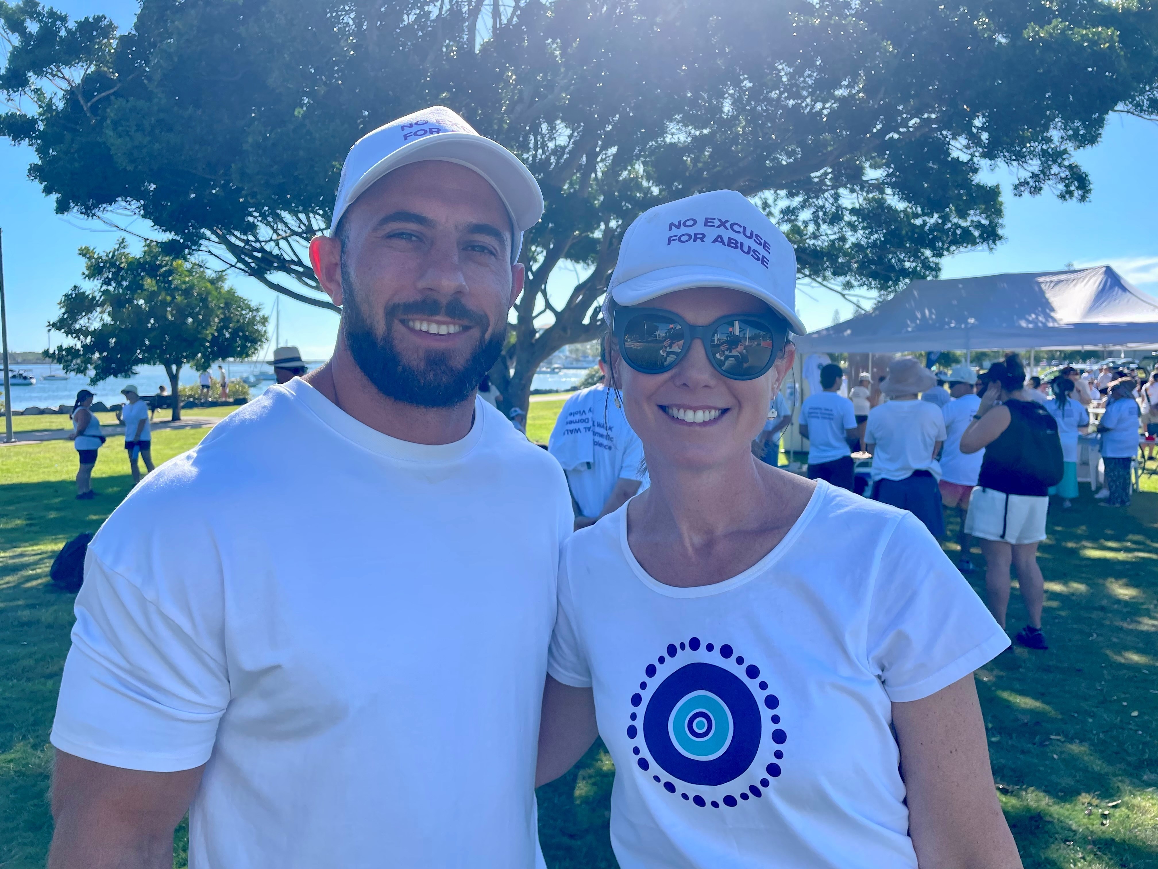 A young man and woman both wearing white shirts and white caps stand together smiling, in a park.