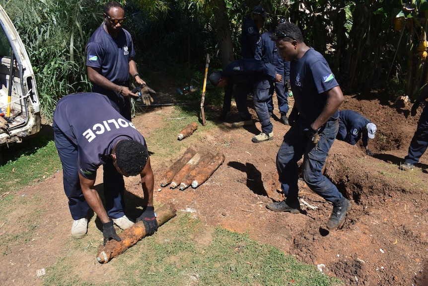 Men in police uniform retrieve old ammunition shells from hole, place them on ground with black gloves.