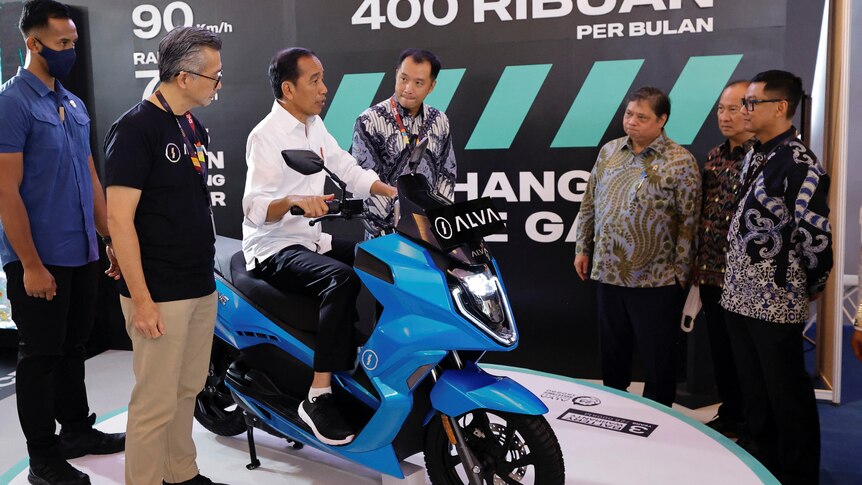 A man sits on a bright blue motorcycle, as several other men watch on.
