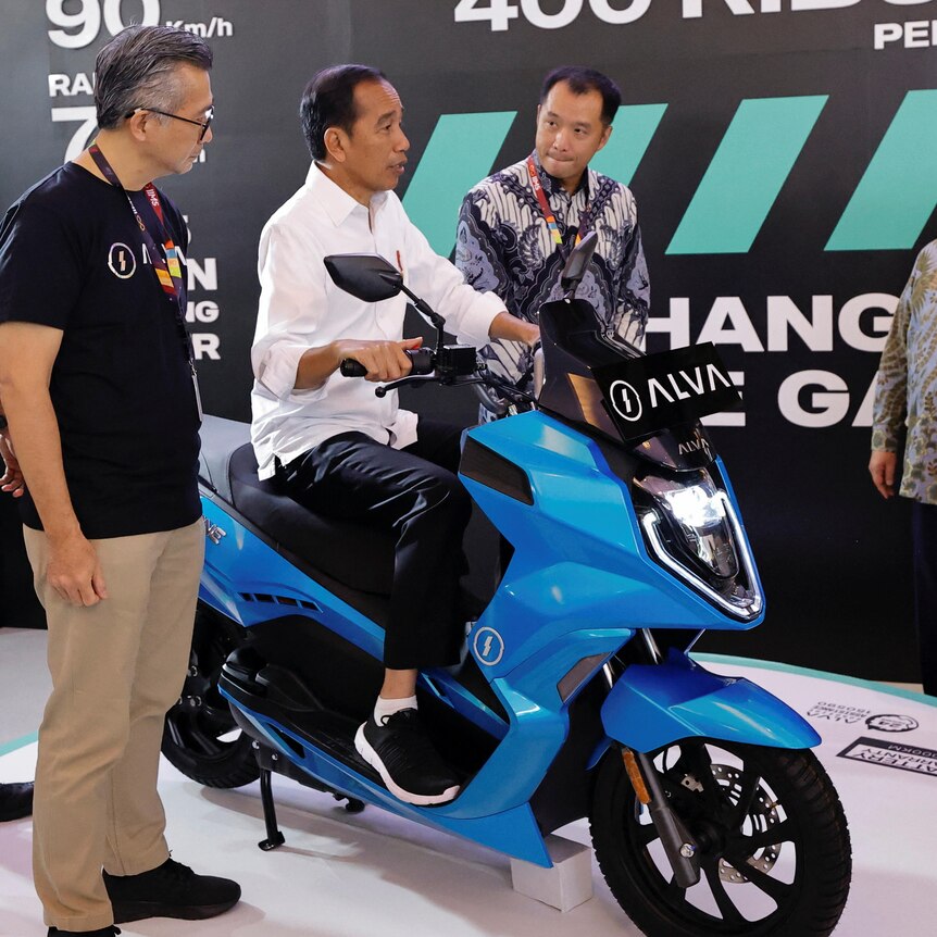 A man sits on a bright blue motorcycle, as several other men watch on.