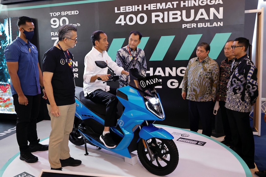 A man sits on a bright blue motorcycle, as several other men watch on.