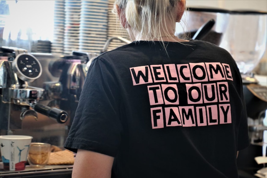 A photo of the back of a t-shirt worn by a woman with blonde hair standing in front of a coffee machine.