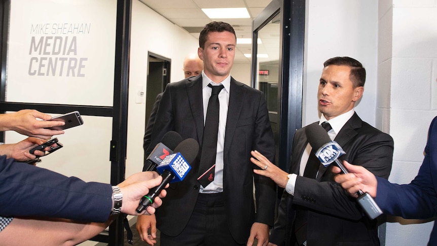 Essendon player Conor McKenna, wearing a suit, walks towards a group of media members with microphones at the AFL tribunal.