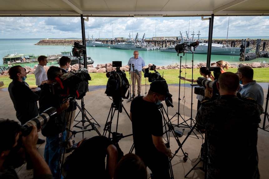 Scott Morrison stands infront of a pack of TV cameras, microphones and journalists with a Navy boat in the background