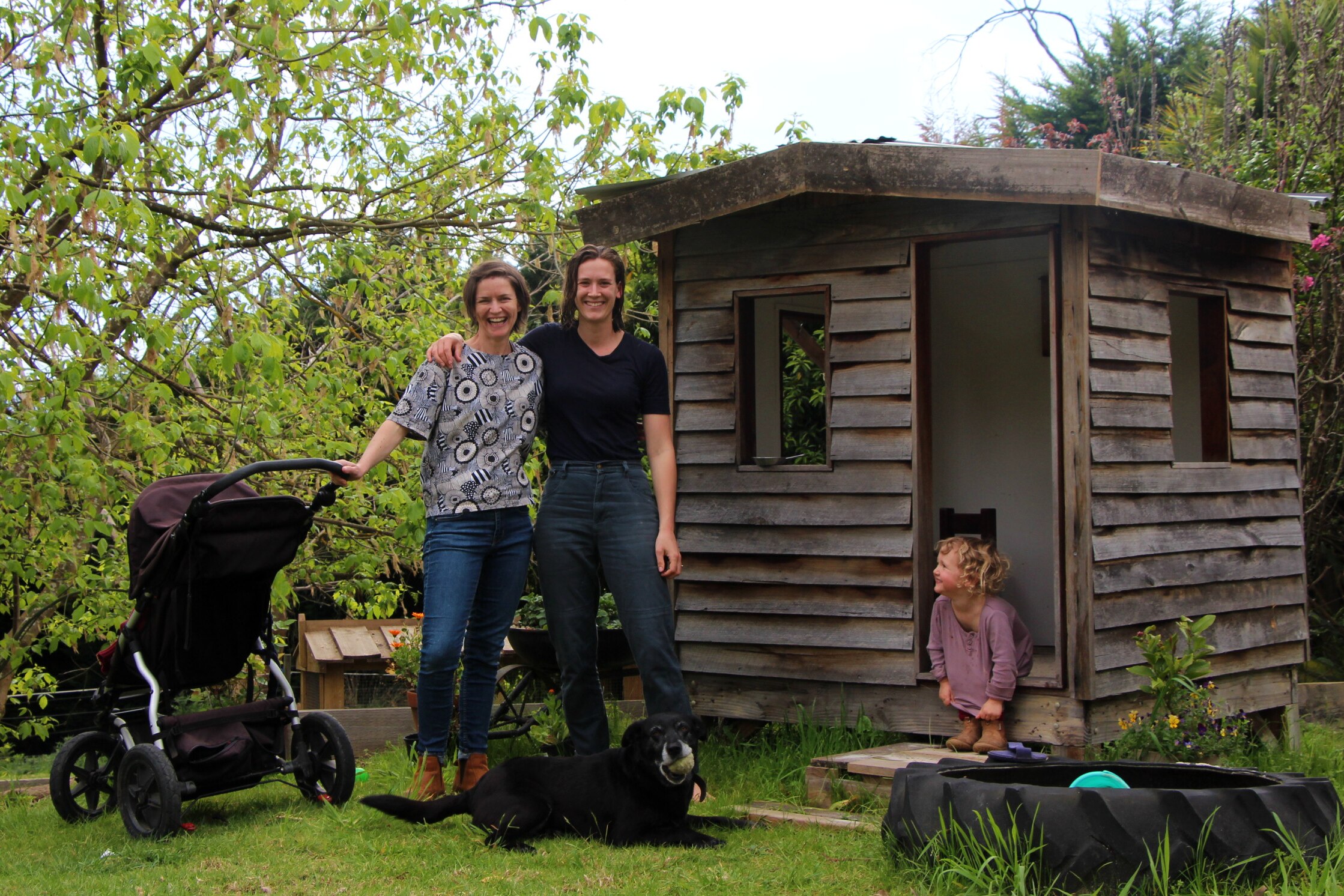 Two women stand with arms around each others shoulders while a two year old girl looks at them smiling
