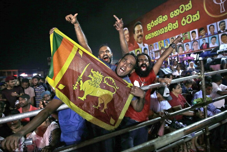 Three men are pictured mid-chant behind a large Sri Lankan flag as they raise their arms behind a crowd barricade.