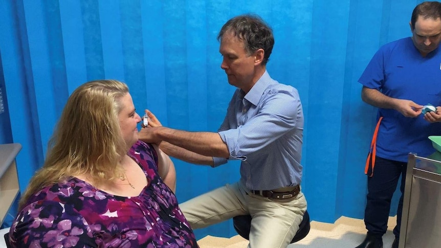 A blonde woman sits down in front of a man who administers a needle into her arm