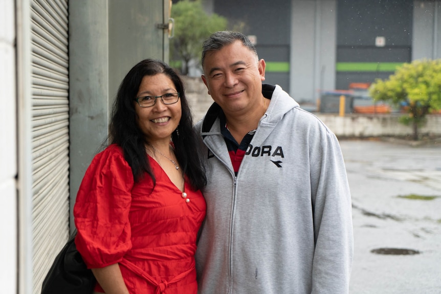 Manuela Jong smiles widely, wearing glasses and red shirt, standing close to man wearing grey jumper, also smiling.