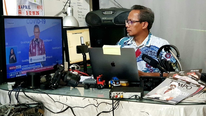 Wu sitting at desk surrounded by microphone, laptop and camera equipment looking at TV screen with man talking about Covid-19.