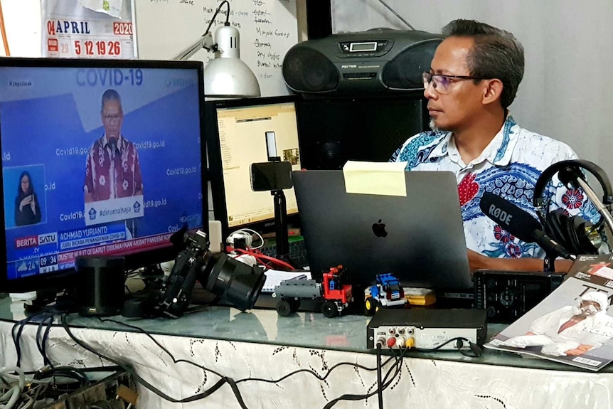 Wu sitting at desk surrounded by microphone, laptop and camera equipment looking at TV screen with man talking about Covid-19.