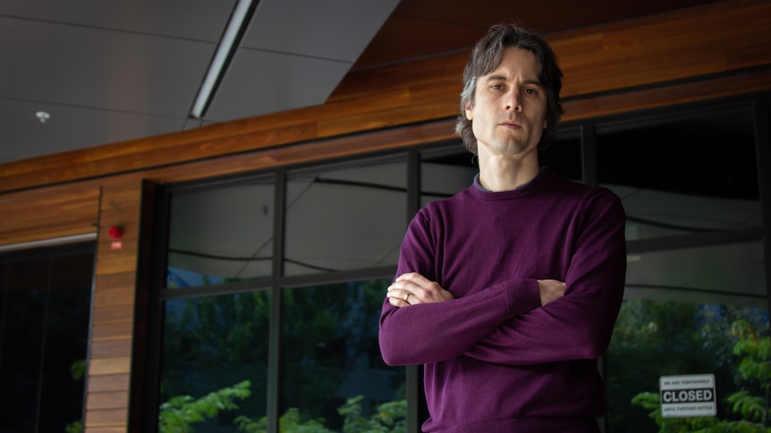 A man with crossed arms stares at the camera, purple jumper