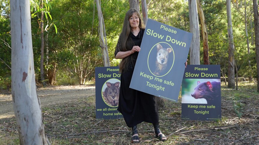 A woman stands in a forest holding a sign saying "Please Slow Down"