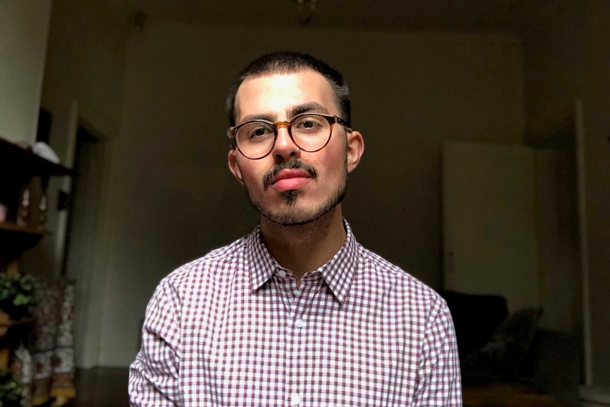 A young man wearing large round glasses, a nose piercing and a collared check shirt.