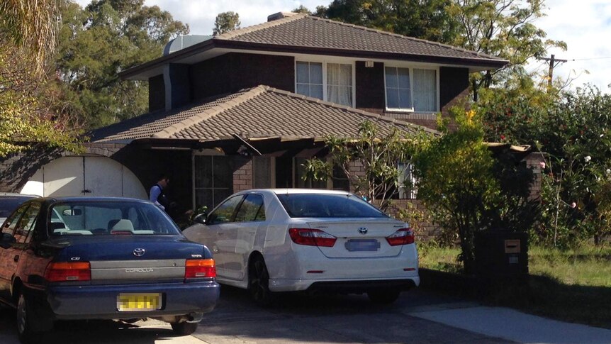 A police officer outside a two-storey brick and tile home. There are two cars parked out the front.