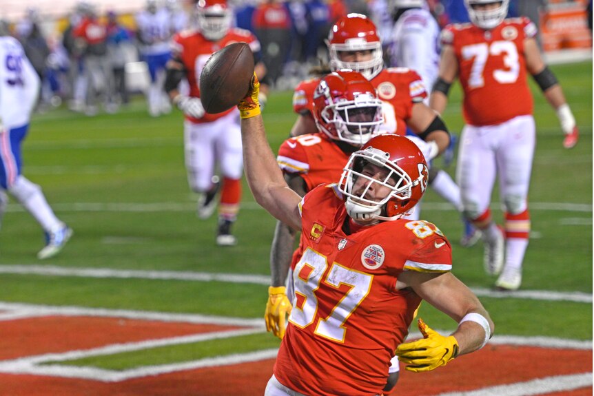 Travis Kelce holds an American football in the air in one hand with his eyes closed