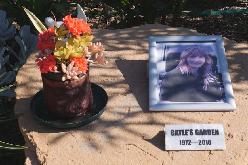 A picture frame with a photo of a blonde-haired woman sits on a rock next to some orange succulent plants.