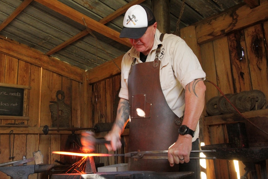 A man in a cap and leather apron, face half hidden by cap, stands over an anvil, hitting a glowing hot knife with a hammer.