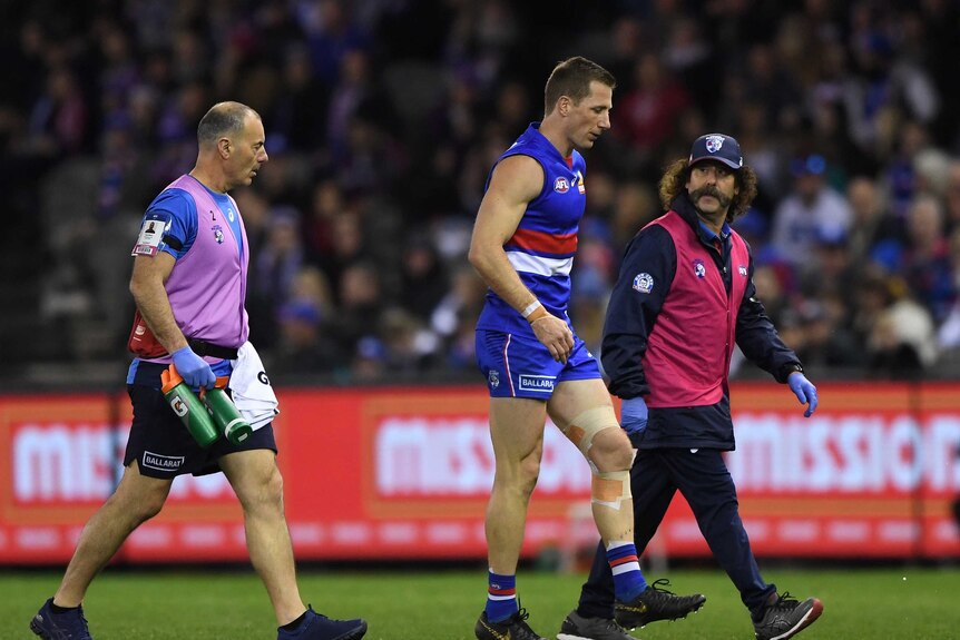 A male AFL player limps off the field with a knee injury while being accompanied by to medical staff members.