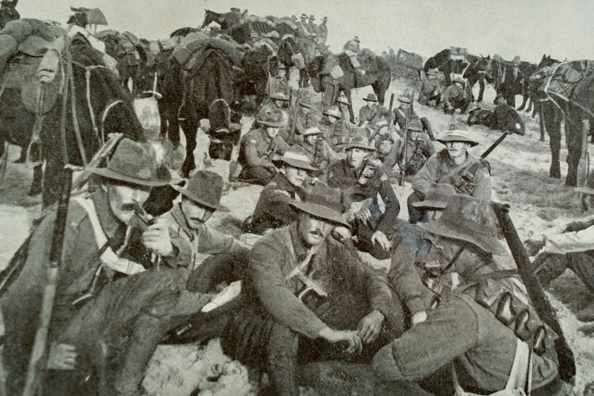 Soldiers of the Australian Light Horse Brigade in Palestine sit on ground with horses in background
