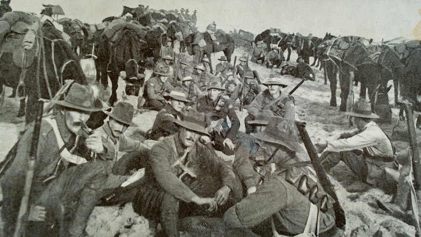 Soldiers of the Australian Light Horse Brigade in Palestine sit on ground with horses in background