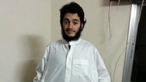 Man with black hair and beard wears white gown stands in carpeted room