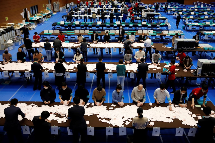A large room with blue carpet shows election officials counting ballots on tables 