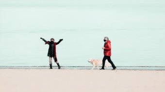 Two people in masks and winter coats walk on a beach with a golden retriever.