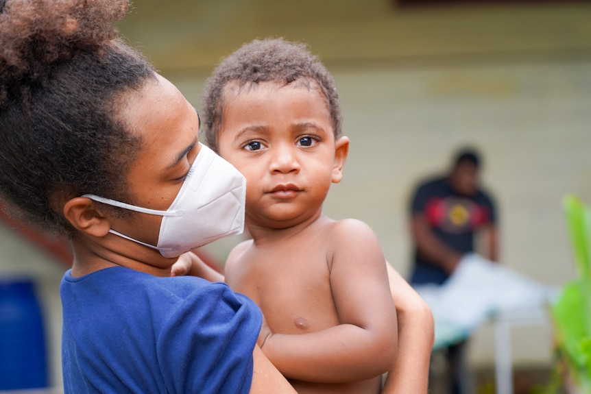 A young woman in a face mask cradles a small child 