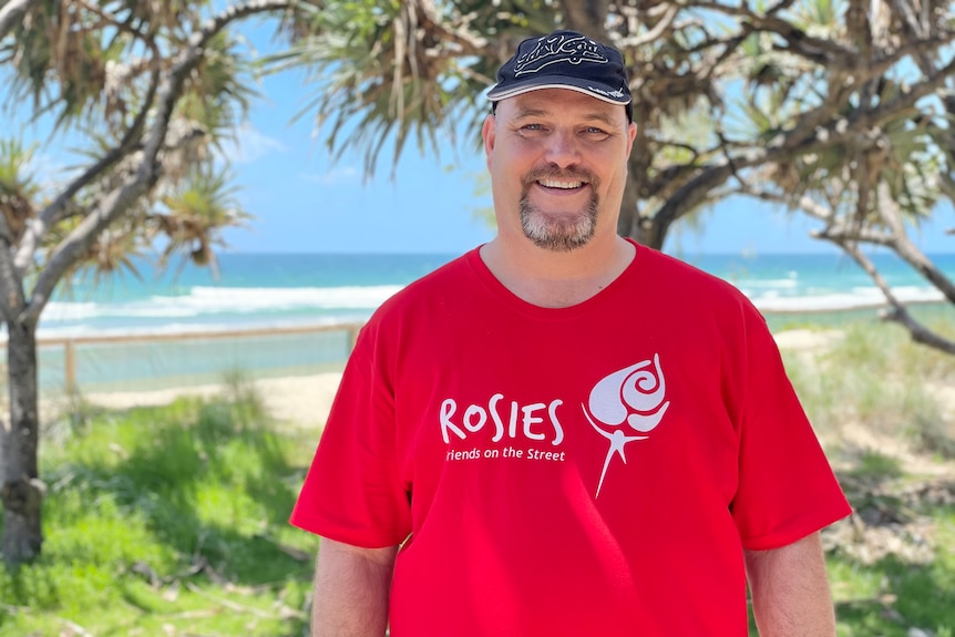 Man wearing cap standing at beach wearing a red shirt that reads "Rosies" with a depiction of a rose