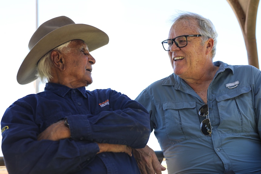An elderly Indigenous man with arms crossed, wearing an Akubra sits next to a middle-aged man wearing glasses.