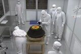 A group of people in white full-body hazmat suits stand around a dark grey capsule in a sterile room.