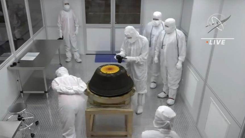 A group of people in white full-body hazmat suits stand around a dark grey capsule in a sterile room.