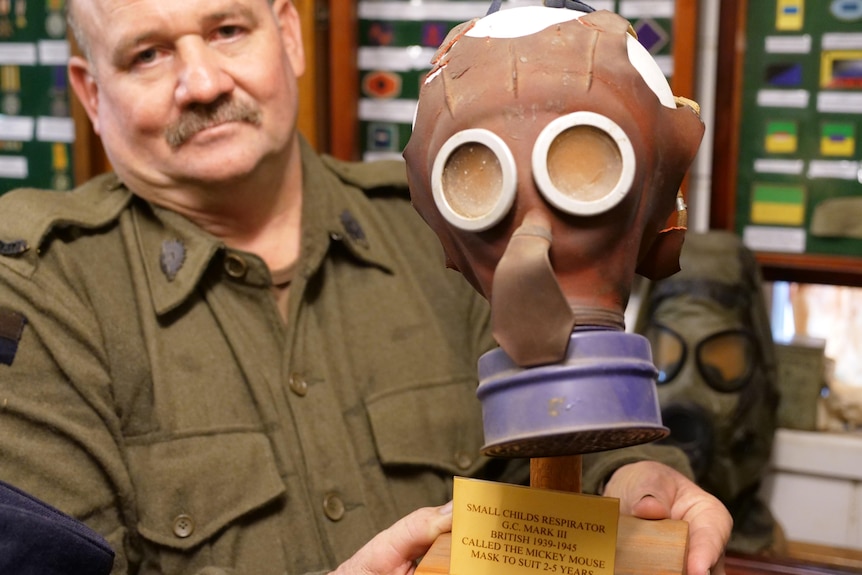 a man dressed in a soldier's outfit holds a brown full face mask built for a small child