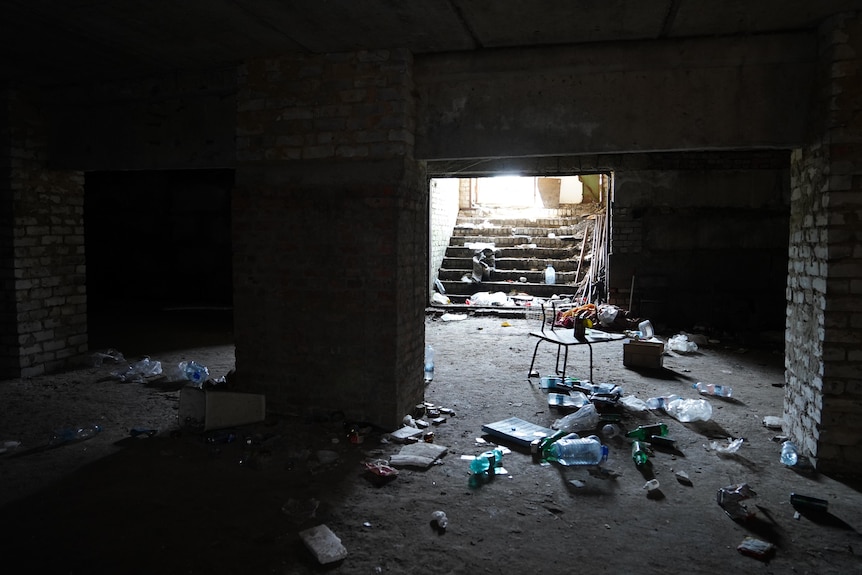 A dark, brick-walled room is littered with debris. Daylight shines in from the top of a flight of stairs descending to the room