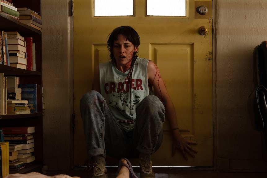 A film still of Kristen Stewart, in 80s-style clothing, crouching with her back against a door. She is covered in blood.