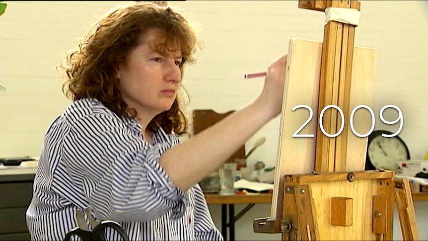 A middle-aged woman with curly brown hair sits at an easel, holding a paintbrush, with a concentrating expression