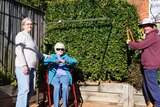 An elderly woman sits on a walker in a backyard with a younger woman while an older man holds garden shears