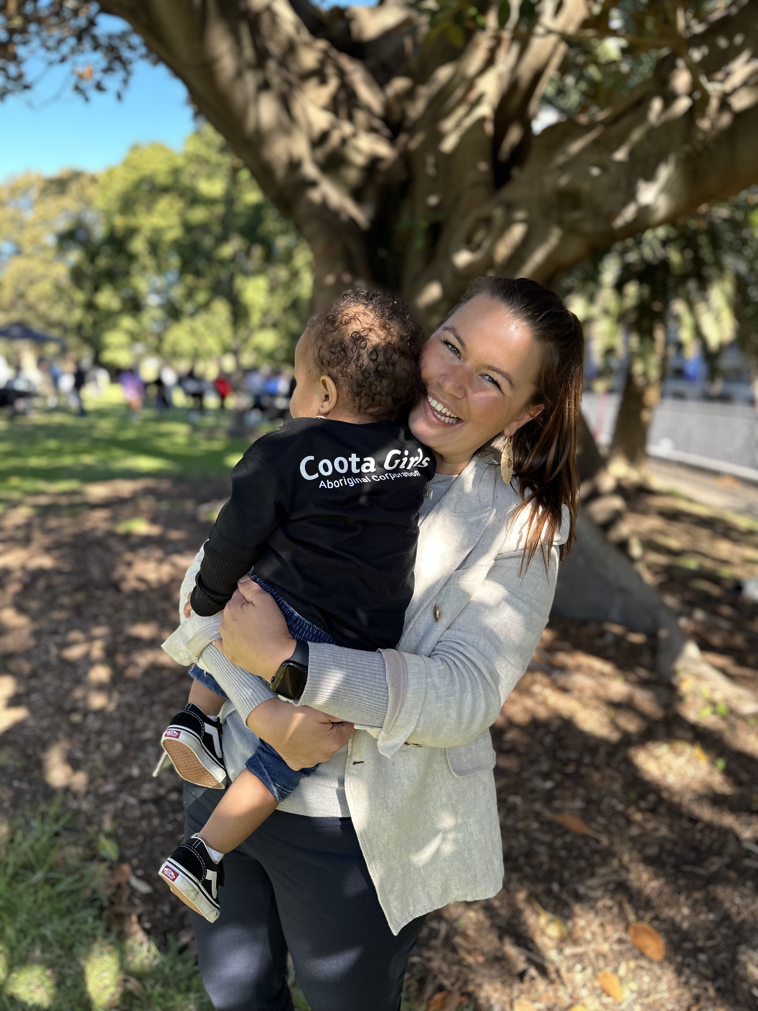 A young Aboriginal woman smiles as she carries a child at an outside event in a green park.