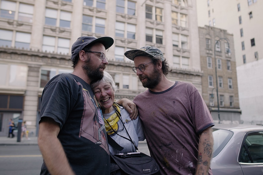 Two bearded men wearing caps and glasses embrace a smiling white-haired woman in a city street.
