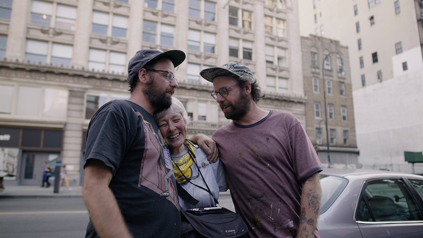 Two bearded men wearing caps and glasses embrace a smiling white-haired woman in a city street.