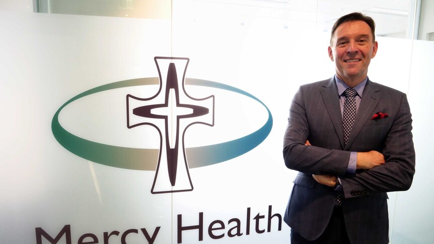 A man dressed in a suit stands in front of a window with a Mercy Health sign