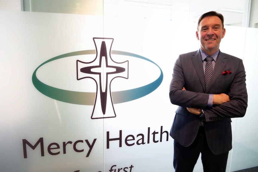 A man dressed in a suit stands in front of a window with a Mercy Health sign