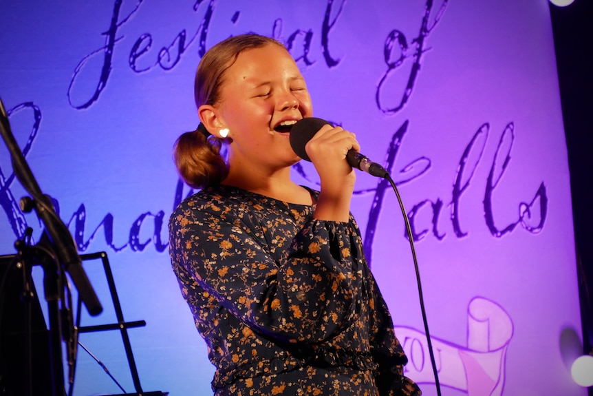 An 11-year-old girl sings into a microphone in front of a Festival of Small Halls banner