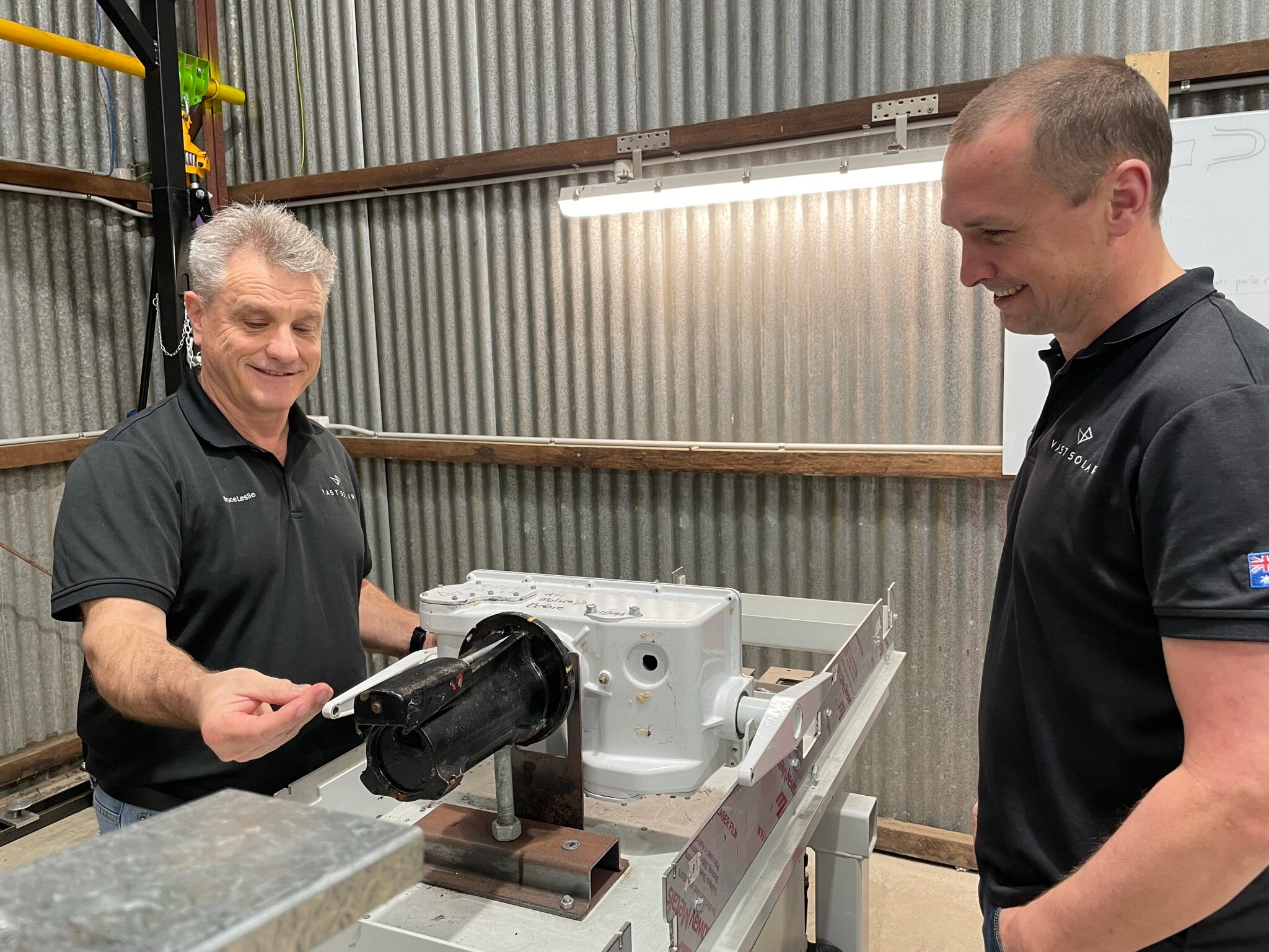 Two men stand near each other in a workshop looking at a machine on a bench.
