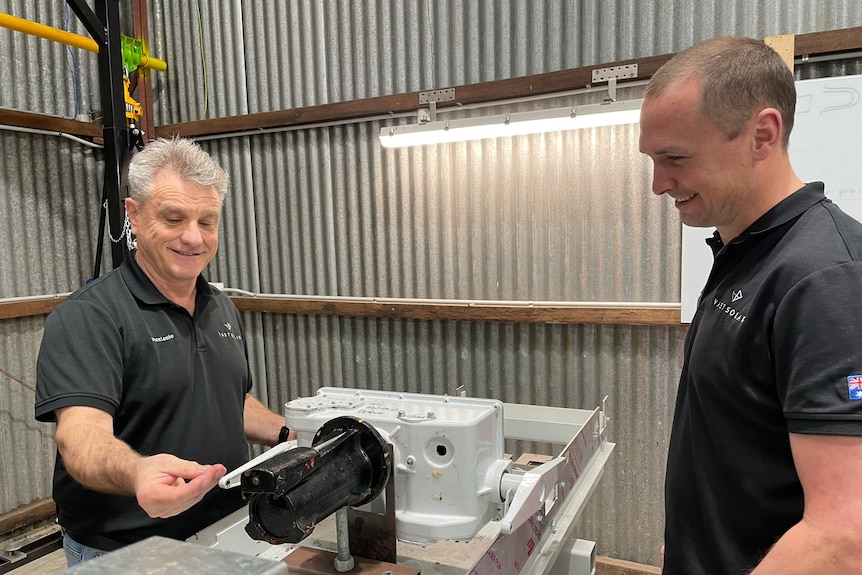 Two men stand near each other in a workshop looking at a machine on a bench.