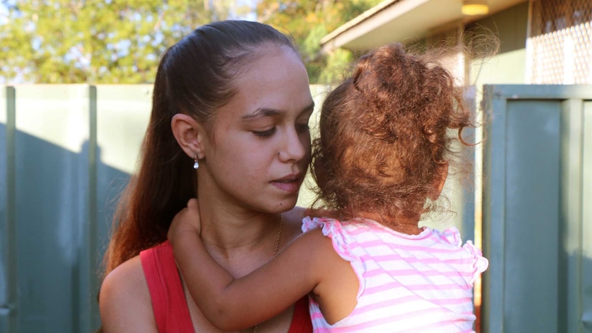 Caitlyn Roe stands outside her house in Broome, looking down, while her child holds onto her neck, looking away from the camera.