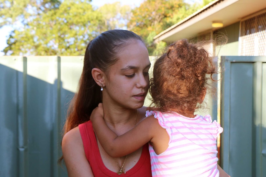 Caitlyn Roe stands outside her house in Broome, looking down, while her child holds unto her neck, looking away from the camera.