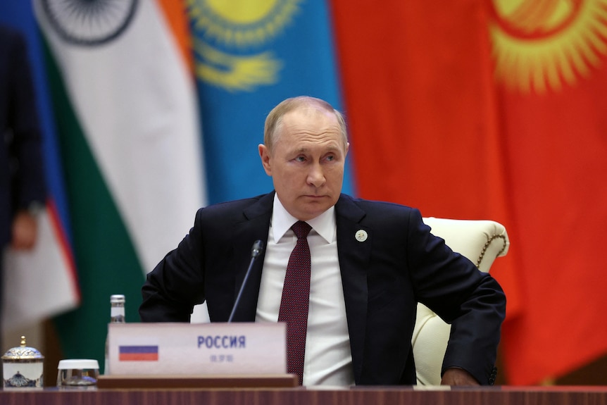 A balding white man wearing a suit sits in a plush-looking chair behind a wooden desk in front of a series of flags.